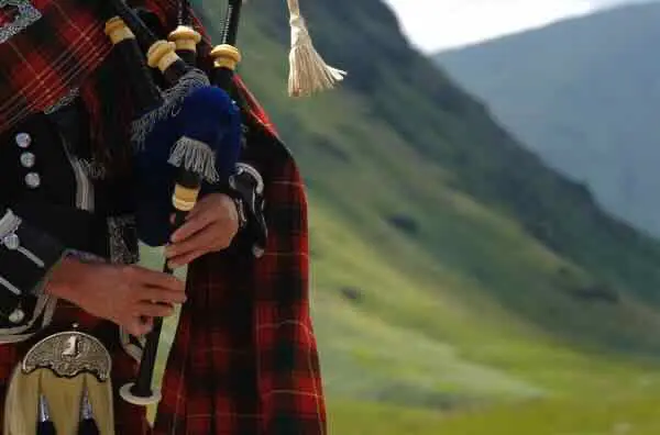 A tartan-clad piper playing bagpipes in a Highland landscape